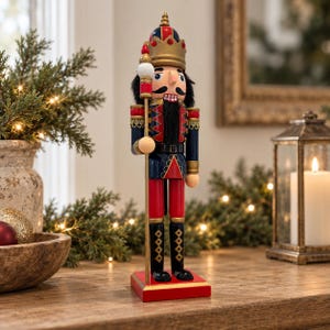 Puede incluir: Una figura de cascanueces de madera sobre una base rectangular roja. El personaje lleva una corona dorada, un uniforme azul y rojo con adornos dorados y botas negras. Sostiene un bastón alto rematado con una esfera blanca sobre una base roja.