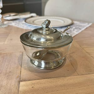 Peut inclure: Un sucrier en argent avec une base en verre et un couvercle orné d'un motif de feuille. Le bol est sur une surface en bois.