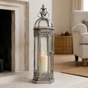 May include: A tall, hexagonal metal lantern with a distressed silver finish, clear glass panels, and a decorative arched top handle. The lantern features intricate metalwork detailing at the base and top, housing a single lit white pillar candle inside.