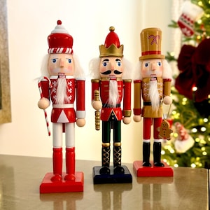 May include: Three decorative wooden nutcrackers in festive attire. The nutcrackers are red, gold, and white, with detailed faces and accessories, including a candy cane and a gingerbread man. They stand on red, blue, and red bases.