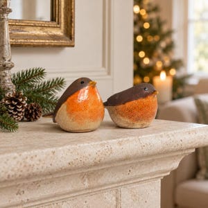 May include: Two ceramic bird figurines featuring dark brown heads and backs with bright orange chests and bellies. Each bird has a small pointed beak and a single black eye. The figurines are rounded in shape and have a smooth, glossy finish.