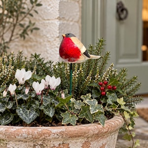 May include: A decorative garden stake featuring a sculpted bird with a bright red chest, a black head, and a cream-colored back. The bird is mounted on a thin green metal rod, designed to be inserted into soil or a planter for outdoor display.