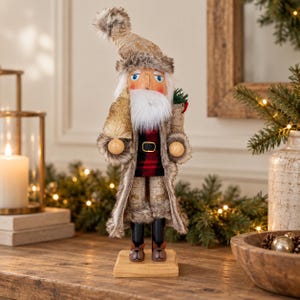 May include: A nutcracker figurine wearing a tan textured hat with faux fur trim, a long tan coat with faux fur lining, and a red and black plaid shirt. The figure has a white beard, holds a small bottle brush tree, and wears brown slippers.