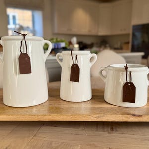 Może przedstawiać: Trzy białe ceramiczne wazy o różnych rozmiarach z uchwytami, każda ozdobiona brązową etykietą. Wazy są ułożone na drewnianej tacy, prezentując minimalistyczny design. Tło to rozmyta sceneria kuchenna.