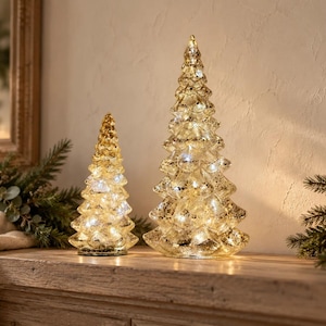 Può includere: Due alberi decorativi conici realizzati in vetro mercurio con una finitura oro metallizzato. Il vetro presenta una consistenza maculata e invecchiata, illuminata dall'interno da piccole luci LED bianco caldo per un effetto luminoso.