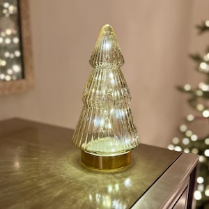 Puede incluir: Un árbol de Navidad decorativo de cristal con una superficie texturizada. El árbol está iluminado con pequeñas luces blancas y se asienta sobre una base dorada. El árbol mide aproximadamente 20 cm de altura.