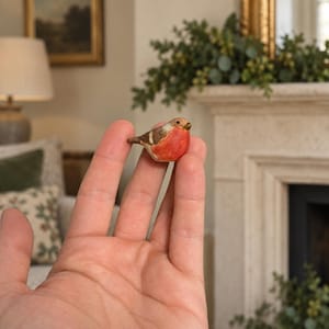 May include: A miniature figurine of a robin bird with a bright orange-red chest, brown wings, and a small dark beak. The bird is crafted with a textured finish and is shown in a scale that fits within the palm of a hand, displaying detailed markings.