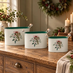 May include: A set of three oval metal containers in graduated sizes, featuring a cream-colored exterior with a green interior rim. Each container displays a painted illustration of pine branches, holly leaves, and red berries tied with a red bow.
