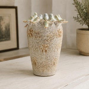 May include: A ceramic vase with a cream-colored base featuring an intricate, lace-like textured pattern in brown. The rim is adorned with a series of small, sculpted bird figures in light blue and green tones, encircling the top opening.