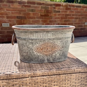 May include: A galvanized metal planter with a distressed finish. The oval-shaped container has two handles and a decorative emblem that reads "FLOWERS & GARDEN". The planter is designed for outdoor use and measures approximately 12 inches long.