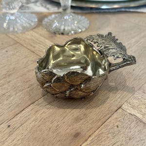 Puede incluir: Un plato de plata con forma de alcachofa con un asa en forma de hoja. El plato es de metal y tiene un acabado brillante.