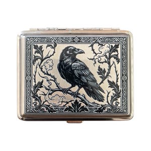 Könnte beinhalten: Eine silberne Zigarettenetui mit einer detaillierten schwarzen Rabenillustration auf einem Ast. Der Rabe ist von einem dekorativen schwarzen Blumenrand auf hellem Hintergrund umgeben. Die Etui hat eine rechteckige Form.
