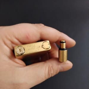 May include: A gold-colored lighter and its component parts are displayed against a dark background. The lighter is rectangular with rounded edges and two circular openings. A separate cylindrical piece with a threaded design is also visible.