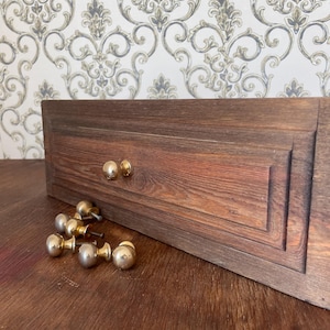 May include: A wooden drawer front with a rich, reddish-brown finish, featuring two installed gold-toned knobs and several loose knobs. The drawer has a recessed panel design. The background is a patterned wallpaper.