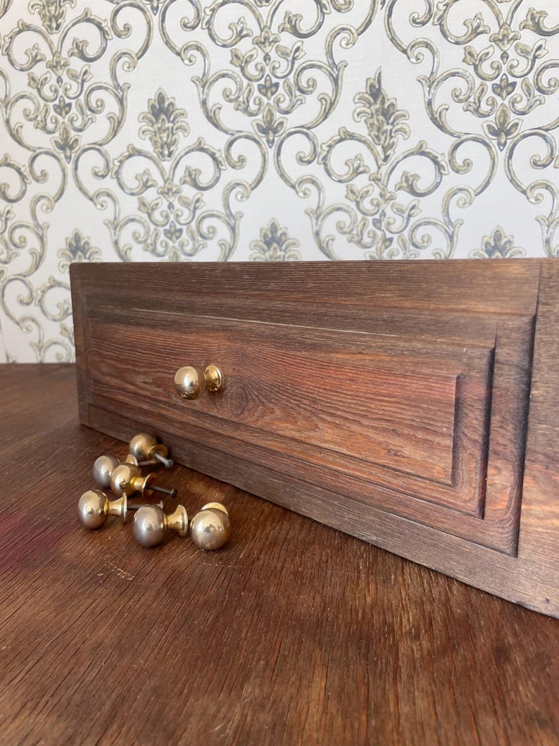 May include: A wooden drawer front with a rich, reddish-brown finish, featuring two installed gold-toned knobs and several loose knobs. The drawer has a recessed panel design. The background is a patterned wallpaper.