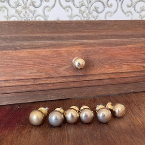 May include: A collection of gold-colored, spherical cabinet knobs and a single knob attached to a wooden drawer. The knobs are arranged on a wooden surface, with a patterned wallpaper in the background.