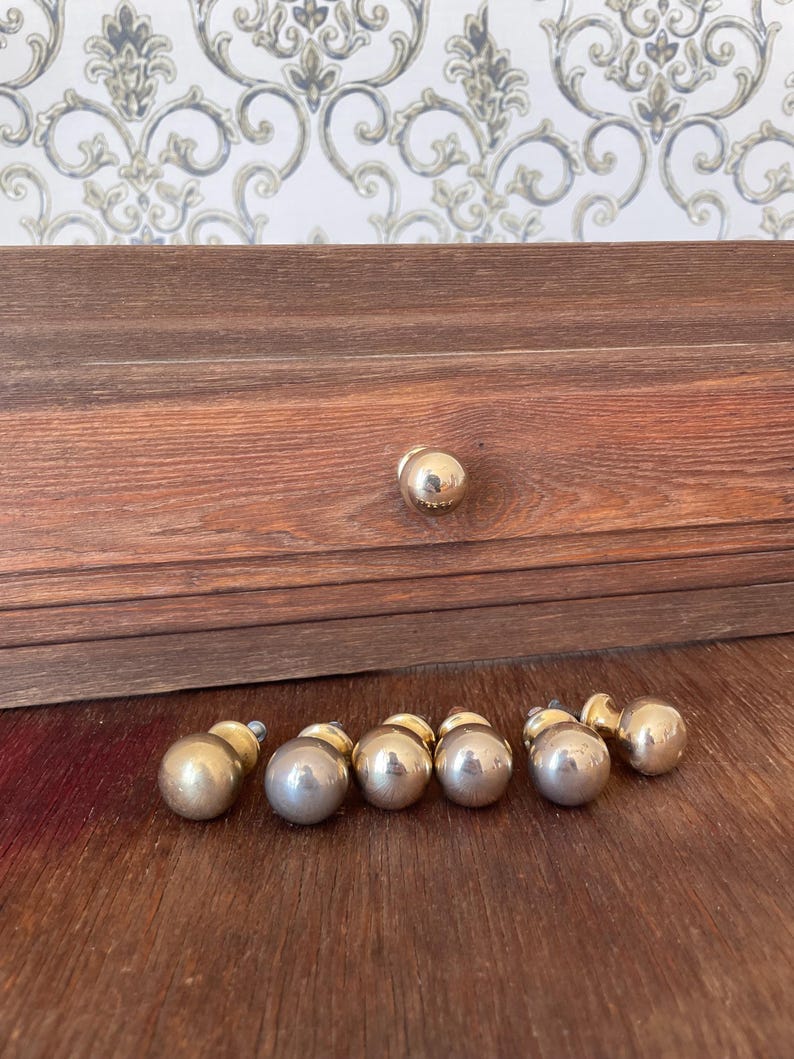 May include: A collection of gold-colored, spherical cabinet knobs and a single knob attached to a wooden drawer. The knobs are arranged on a wooden surface, with a patterned wallpaper in the background.