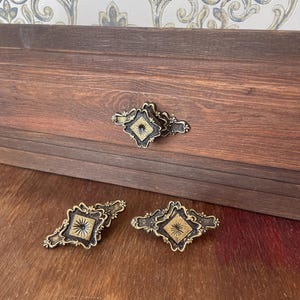 May include: Three ornate metal drawer pulls featuring a diamond-shaped center with a sunburst pattern, finished in a dark antique bronze tone with gold-colored highlights. One pull is attached to a wooden drawer front, while two lie flat.
