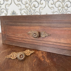 Puede incluir: Primer plano de un cajón de madera con dos tiradores antiguos de latón. Los tiradores tienen un diseño floral y están montados sobre una superficie de madera marrón oscuro. El fondo presenta un papel pintado con motivos.