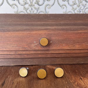 Pode incluir: Um conjunto de quatro puxadores circulares para armários, cada um com um anel exterior de madeira castanha escura e um centro plano de metal dourado escovado. Um puxador está fixado numa frente de gaveta de madeira escura, três estão alinhados.