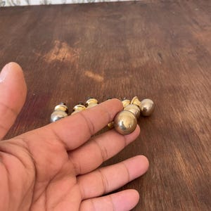 May include: A collection of small, round, metallic knobs with a gold and silver finish. The knobs are arranged on a dark wooden surface. One knob is held in a hand, showcasing its spherical shape and reflective surface.