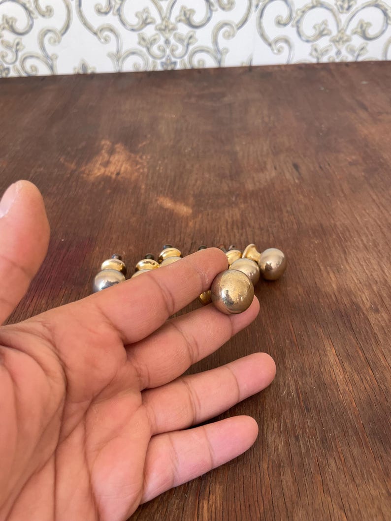 May include: A collection of small, round, metallic knobs with a gold and silver finish. The knobs are arranged on a dark wooden surface. One knob is held in a hand, showcasing its spherical shape and reflective surface.