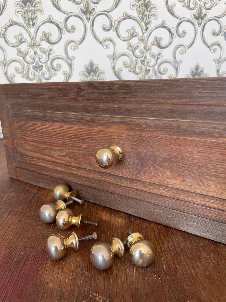 May include: A collection of brass cabinet knobs with a polished finish, some attached to a dark brown wooden drawer. The knobs are round and have a classic design. The wood grain is visible on the drawer.