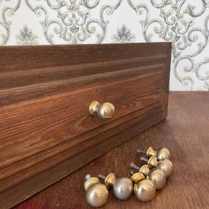May include: A wooden drawer with two gold-colored knobs and a collection of similar knobs. The drawer is made of dark brown wood. The knobs are round and shiny. The background features a patterned wallpaper.