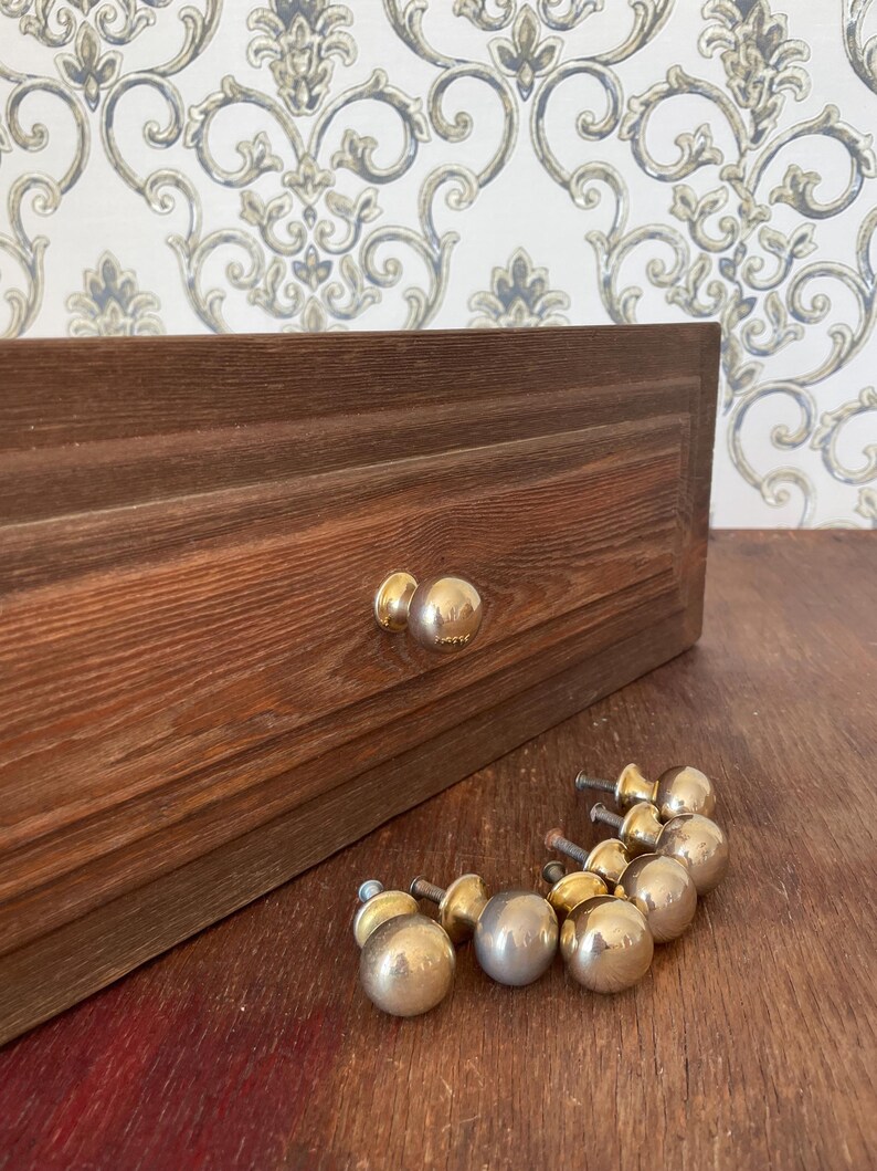 May include: A wooden drawer with two gold-colored knobs and a collection of similar knobs. The drawer is made of dark brown wood. The knobs are round and shiny. The background features a patterned wallpaper.