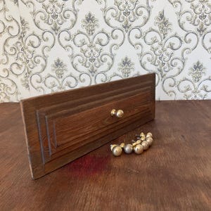 May include: A wooden drawer front with two gold knobs and a collection of gold-colored cabinet knobs. The drawer front is a dark brown color with a rectangular shape and a carved design. The knobs are round.