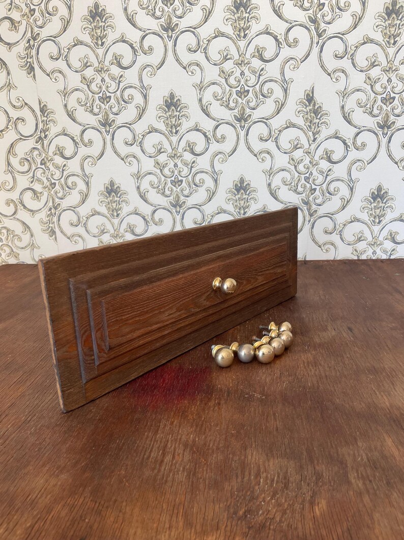 May include: A wooden drawer front with two gold knobs and a collection of gold-colored cabinet knobs. The drawer front is a dark brown color with a rectangular shape and a carved design. The knobs are round.