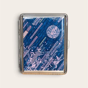 May include: A rectangular silver metal cigarette case featuring a glossy top panel with a dark blue background. The design displays a pale pink full moon, abstract geometric lines, and stylized floral patterns in a light pink hue.