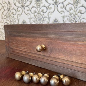 May include: A wooden drawer with two brass knobs and a collection of brass ball knobs. The drawer is made of dark brown wood with visible grain. The knobs are round and shiny, suggesting a vintage or antique style.
