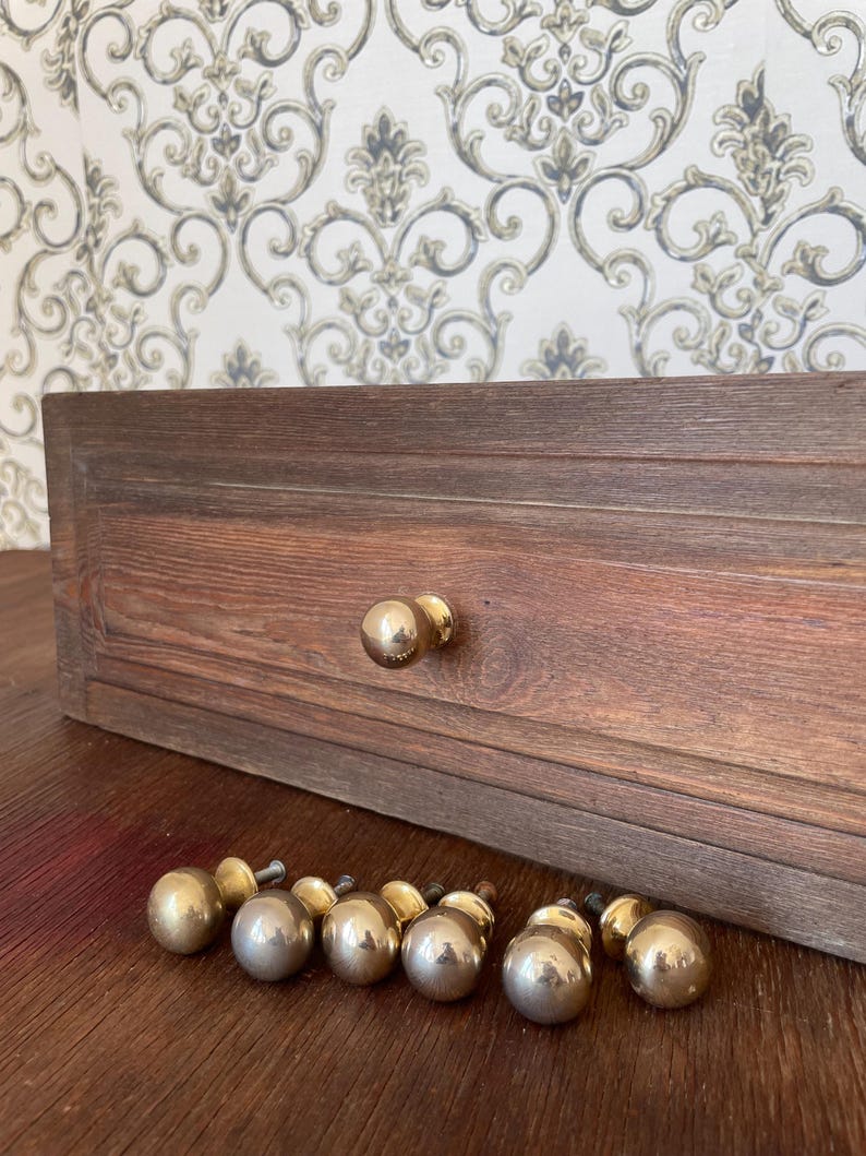 May include: A wooden drawer with two brass knobs and a collection of brass ball knobs. The drawer is made of dark brown wood with visible grain. The knobs are round and shiny, suggesting a vintage or antique style.