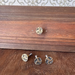 Puede incluir: Cuatro pomos circulares de latón para armarios con un patrón de relieve texturizado y giratorio. Un pomo está fijado a un frente de cajón de madera, mientras que tres pomos idénticos están colocados delante sobre una superficie de madera, cada uno con un tornillo.