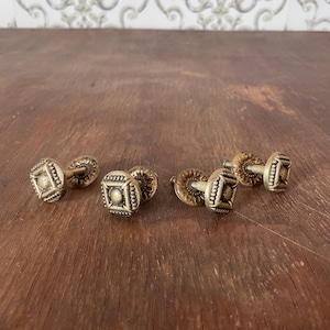 May include: Four antique brass-toned metal cabinet knobs featuring a square face with a central circular detail and a decorative, scalloped circular base plate. Each knob includes a threaded metal screw extending from the back for installation.