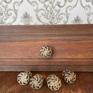Puede incluir: Un conjunto de seis tiradores decorativos para armarios. Los tiradores tienen un diseño en espiral con un acabado dorado y detalles en marrón oscuro. Un tirador está fijado a un cajón de madera, los otros están sobre una superficie de madera.