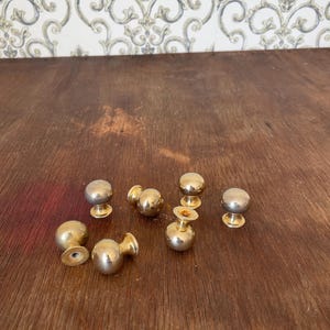 May include: A collection of seven brass cabinet knobs with a rounded design. The knobs have a shiny, metallic finish and are arranged on a wooden surface. The knobs are a classic hardware choice.
