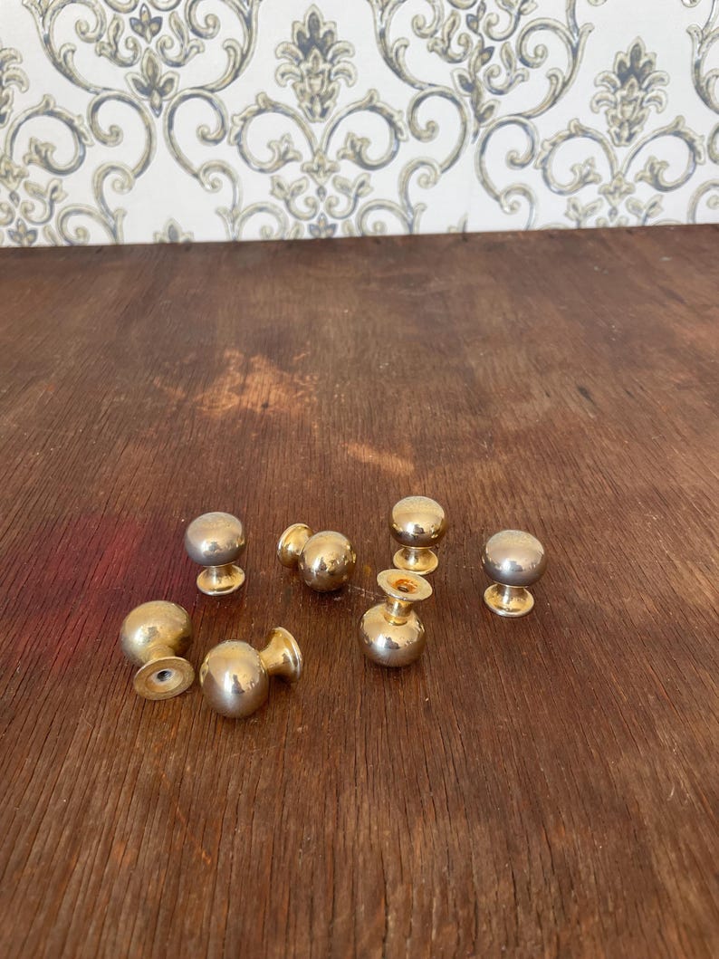 May include: A collection of seven brass cabinet knobs with a rounded design. The knobs have a shiny, metallic finish and are arranged on a wooden surface. The knobs are a classic hardware choice.