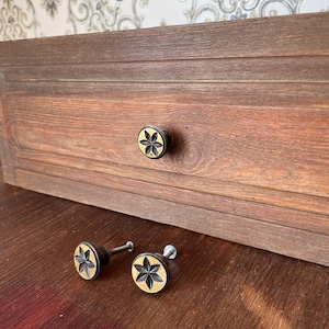 May include: Wooden drawer with a dark finish and a flower-shaped knob. Two additional knobs with the same design are on the wooden surface. The knobs have a black flower design with a gold-colored background.