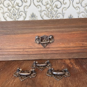May include: Close-up of antique bronze drawer pulls with ornate designs. Three pulls are arranged on a wooden surface, with one attached to a wooden drawer. The pulls have a vintage aesthetic, perfect for furniture restoration projects.