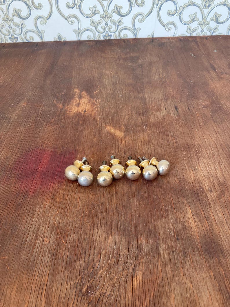 May include: A collection of seven small, round cabinet knobs. Each knob has a silver-colored, spherical body and a gold-colored base. The knobs are arranged on a wooden surface with a warm, brown tone.