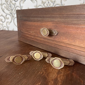 May include: Four brass-toned metal drawer pulls featuring an octagonal center plate with a smooth finish, flanked by textured, ribbed horizontal bars with decorative scrollwork ends. One pull is attached to a wooden drawer front.