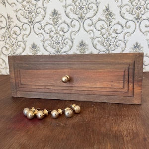 May include: A rectangular, wooden drawer with a single gold knob and a set of six gold knobs. The drawer is a dark brown color and has a recessed panel design. The knobs are round and shiny, contrasting with the wood's texture.