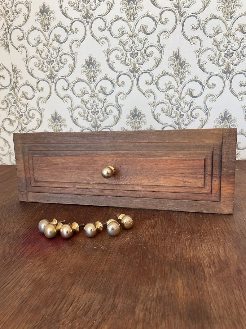 May include: A rectangular, wooden drawer with a single gold knob and a set of six gold knobs. The drawer is a dark brown color and has a recessed panel design. The knobs are round and shiny, contrasting with the wood's texture.