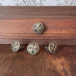 May include: Four round buttons featuring a black center with a raised gold-toned metallic floral pattern and a textured rope-style border. Each button has a shank on the back for attachment, displaying a consistent circular design and metallic finish.