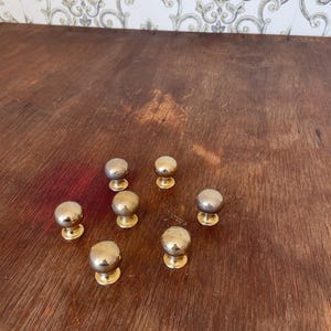 May include: A collection of seven brass cabinet knobs with a rounded top and a circular base. The knobs are arranged on a dark wooden surface. The knobs have a shiny, metallic finish.