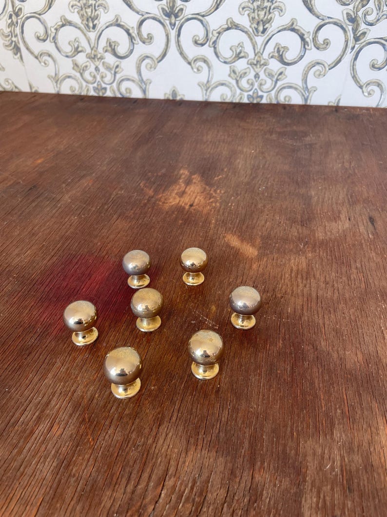 May include: A collection of seven brass cabinet knobs with a rounded top and a circular base. The knobs are arranged on a dark wooden surface. The knobs have a shiny, metallic finish.
