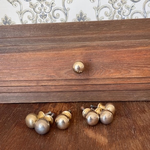 May include: A set of vintage brass cabinet knobs and drawer pulls. The knobs are spherical with a polished finish. The drawer pulls have a similar design, with a small base and a larger spherical top. The wood drawer is visible in the background.
