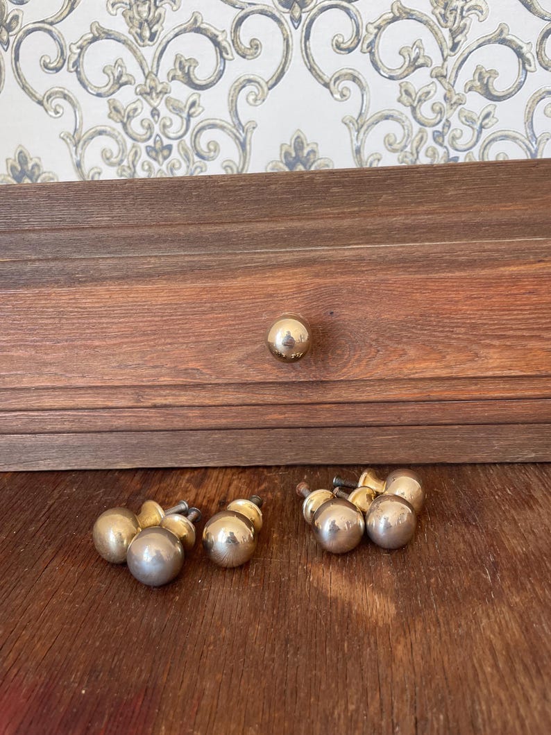 May include: A set of vintage brass cabinet knobs and drawer pulls. The knobs are spherical with a polished finish. The drawer pulls have a similar design, with a small base and a larger spherical top. The wood drawer is visible in the background.